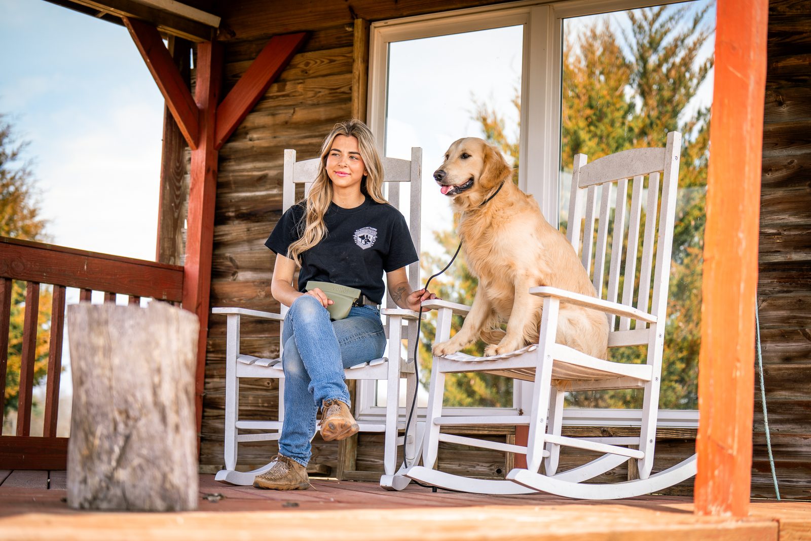 A trainer sits on a log-cabin porch beside a golden retriever in a rocking chair with fall color behind